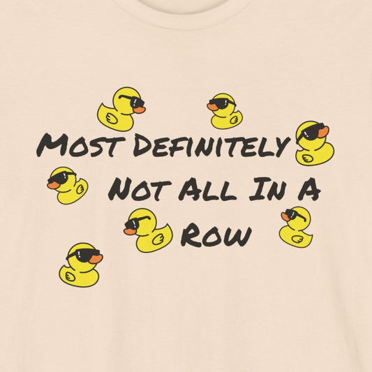 A light beige crewneck t-shirt featuring a graphic print of yellow rubber ducks and the text 'MOST DEFINITELY NOT ALL IN A ROW' in black.