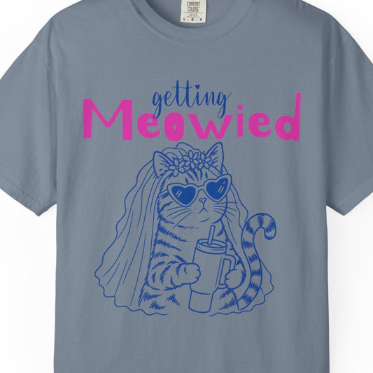A slate blue crewneck t-shirt featuring a graphic of a cat wearing sunglasses and a veil with the text 'getting Meowed' in blue and pink, displayed on a clean white background.
