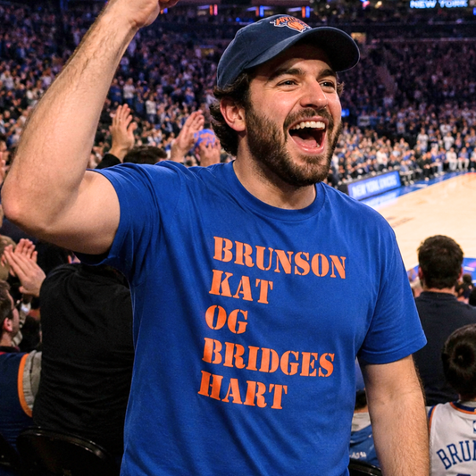 A man in a crowded arena wearing a blue New York Knicks cap and a blue t-shirt with orange text reading 'BRUNSON KAT OG BRIDGES HART', cheering with his arm raised.