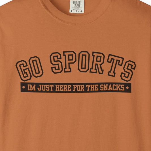 A rust-orange short-sleeve t-shirt with a black graphic on the chest that reads 'GO SPORTS' and 'I'M JUST HERE FOR THE SNACKS'.
