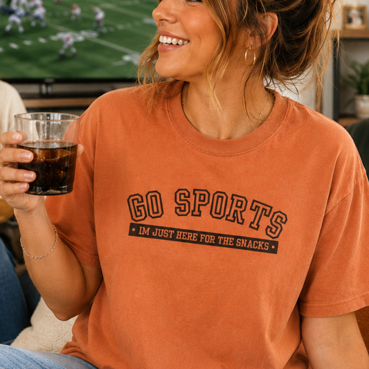 A woman wearing a burnt orange graphic t-shirt with the text 'GO SPORTS' and 'I'M JUST HERE FOR THE SNACKS' printed in black. She is holding a glass of cola in a cozy indoor setting.