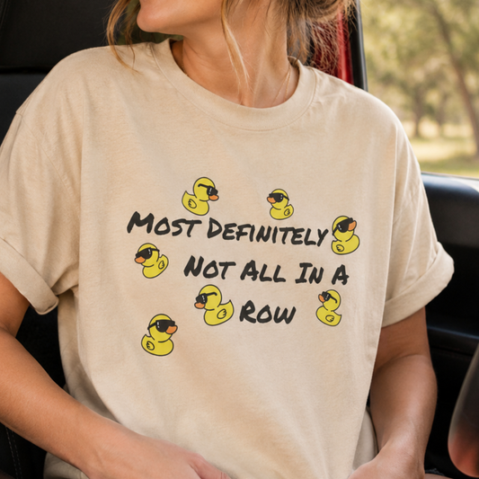 A woman wearing a beige graphic t-shirt with yellow rubber duckies and the text 'MOST DEFINITELY NOT ALL IN A ROW' while sitting in a car.