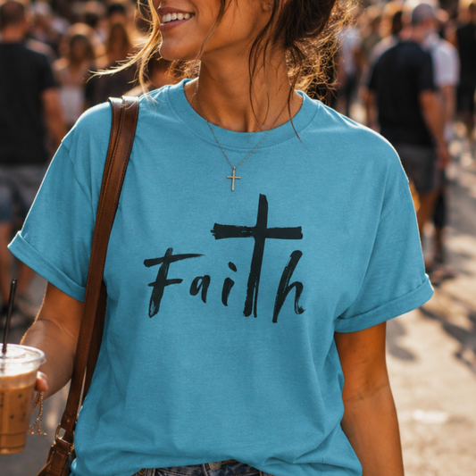 A woman wearing a light blue crewneck t-shirt with the word 'Faith' printed in black script across the chest, featuring a cross as the letter 'i'. She is carrying a brown shoulder bag and holding an iced coffee in a crowded outdoor setting.