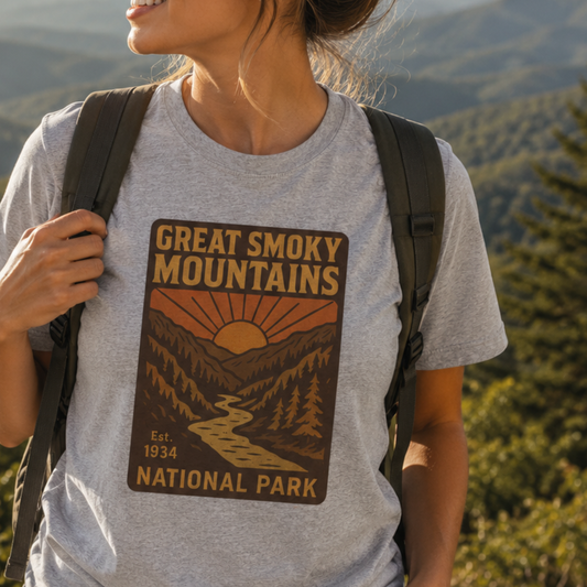 A woman wearing a heather gray t-shirt with a 'Great Smoky Mountains National Park' graphic print, carrying a black backpack outdoors with a blurred mountain forest background.