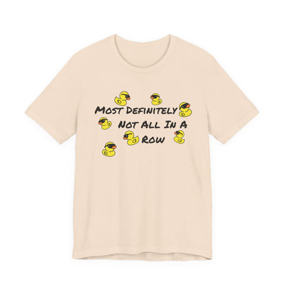 A light beige crewneck t-shirt featuring a graphic print of yellow rubber ducks and the text 'MOST DEFINITELY NOT ALL IN A ROW' in black.