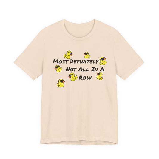 A light beige crewneck t-shirt featuring a graphic print of yellow rubber ducks and the text 'MOST DEFINITELY NOT ALL IN A ROW' in black.