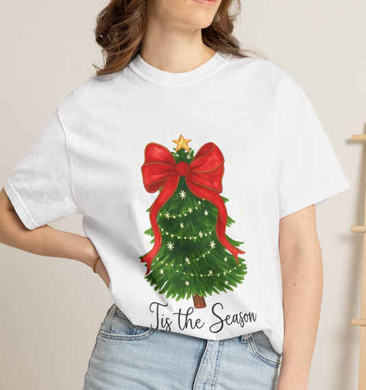 A woman wearing a white graphic t-shirt with a Christmas tree design and the text 'It's the Season', paired with light blue denim shorts in a cozy indoor setting.