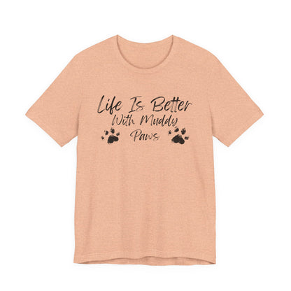 A heather peach short-sleeve t-shirt with a black graphic print that reads 'Life Is Better With Muddy Paws' accompanied by two paw prints, shown on a plain white background.
