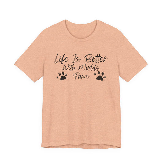 A heather peach short-sleeve t-shirt with a black graphic print that reads 'Life Is Better With Muddy Paws' accompanied by two paw prints, shown on a plain white background.