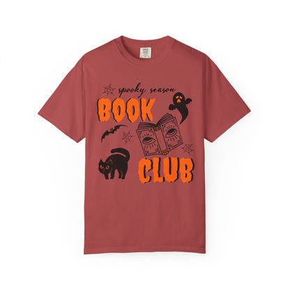 A dusty rose short-sleeve t-shirt with a Halloween-themed graphic that reads 'spooky season BOOK CLUB' in orange and black, featuring illustrations of a black cat, ghost, book, and bat on a white background.