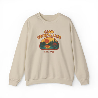 A beige crewneck sweatshirt featuring a retro-style graphic of a campfire scene at Crystal Lake with the text 'CAMP CRYSTAL LAKE' and 'EST. 1935'.