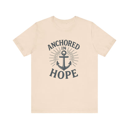 A light beige crewneck t-shirt with a dark navy graphic on the front featuring an anchor and the text 'ANCHORED IN HOPE'.