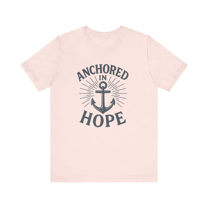 A light pink short-sleeve crewneck t-shirt with a dark gray graphic on the front featuring an anchor and the words 'ANCHORED IN HOPE'.