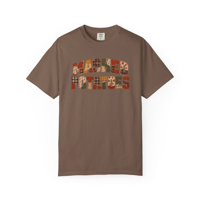 A brown short-sleeve crewneck t-shirt featuring a graphic on the chest that reads 'MASKED PATRIOTS' in a patchwork pattern of autumn colors like orange, red, and brown plaid.