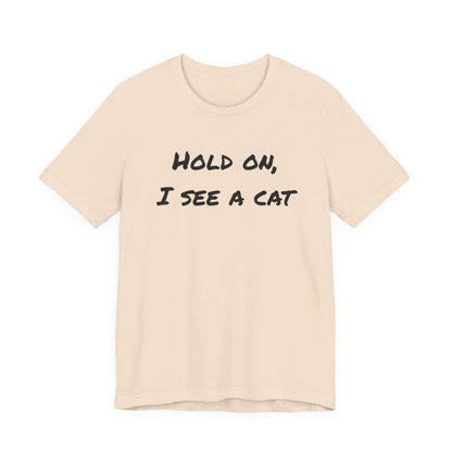 A light beige crewneck t-shirt with the phrase 'HOLD ON, I SEE A CAT' printed in a black handwritten-style font across the chest, displayed on a solid white background.
