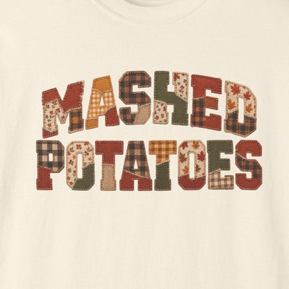 An off-white crewneck t-shirt featuring the words 'MASHED POTATOES' printed across the chest in a rustic, patterned font with shades of brown, orange, and beige.