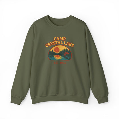 An olive green crewneck sweatshirt featuring a retro-style 'Camp Crystal Lake' graphic on the chest with a sunset, trees, and a deer silhouette.