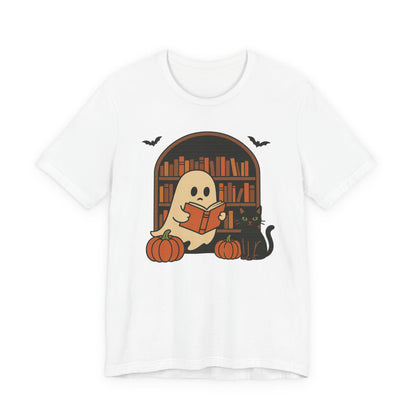 A white short-sleeve crewneck t-shirt featuring a cute Halloween graphic of a ghost reading a book in front of a bookshelf, with pumpkins, a black cat, and bats.