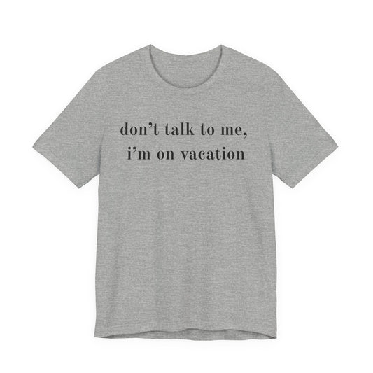 A heather gray short-sleeve t-shirt with the text 'don't talk to me, i'm on vacation' printed in black lowercase serif font across the chest, displayed on a solid white background.