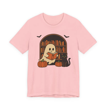 A light pink short-sleeve t-shirt with a cartoon graphic of a ghost reading a book in front of a bookshelf, with pumpkins, a black cat, and bats.