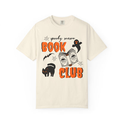 An off-white short-sleeve crewneck t-shirt featuring a 'spooky season BOOK CLUB' graphic with orange text, a cartoon book, a black cat, and a ghost on a plain white background.