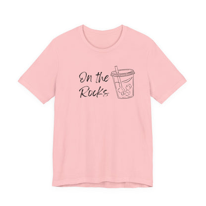 A light pink short-sleeve crewneck t-shirt with a black graphic print on the front that reads 'On the Rocks' next to a simple line drawing of a iced coffee cup, shown against a plain white background.