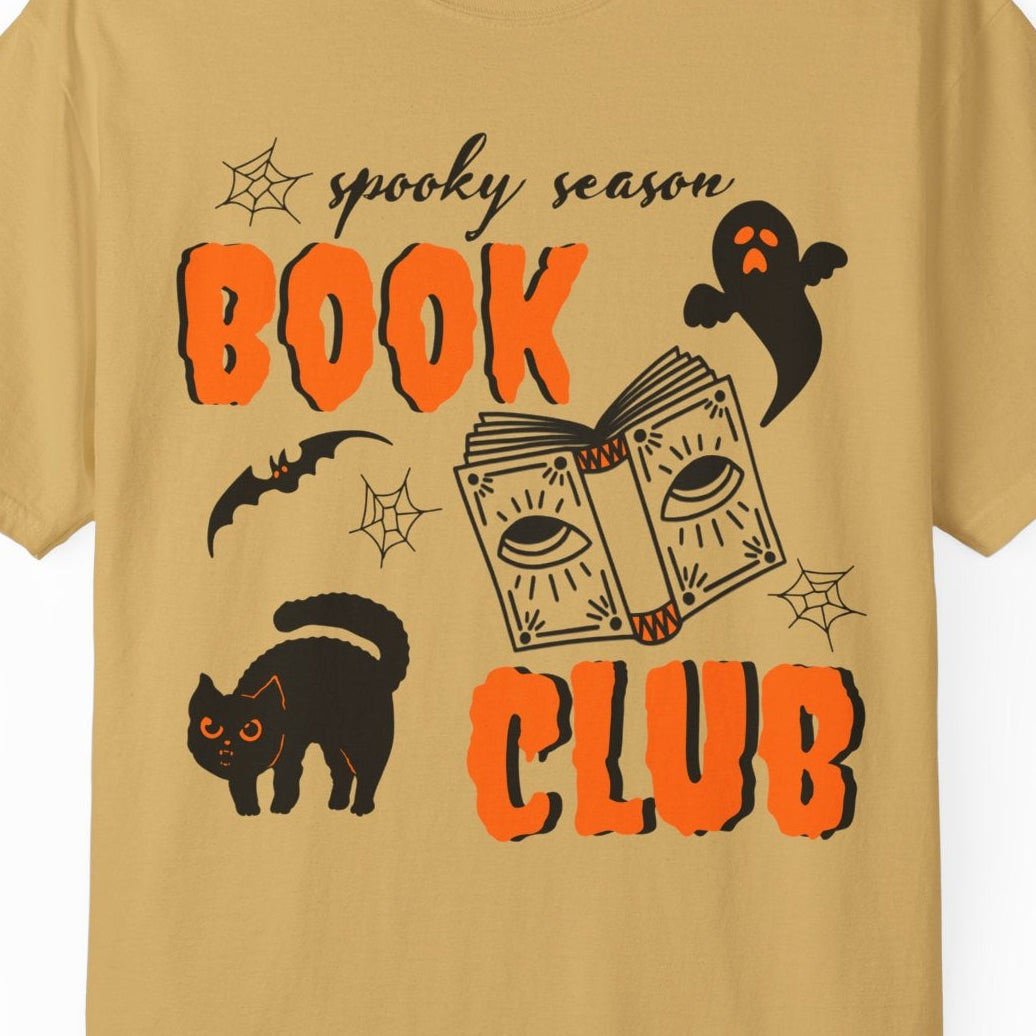 A tan crewneck t-shirt featuring a 'spooky season BOOK CLUB' graphic with Halloween illustrations including a black cat, ghost, bat, and book on a white background.