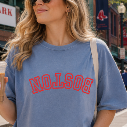 A woman wearing a light blue crewneck t-shirt with 'BOSTON' printed in red arched letters across the chest, holding an iced coffee on a city street.