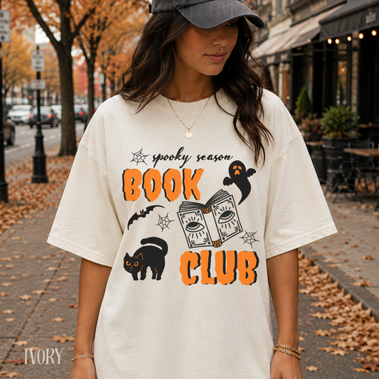 A woman wearing an oversized cream-colored graphic t-shirt with 'spooky season BOOK CLUB' printed in orange and black, standing on a city sidewalk with autumn leaves and trees in the background.