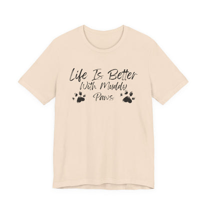 A light beige short-sleeve t-shirt with a black graphic print on the front that reads 'Life Is Better With Muddy Paws' accompanied by two paw prints, shown against a plain white background.