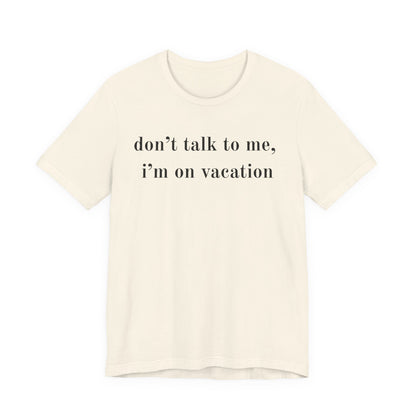 An off-white short-sleeve crewneck t-shirt with the text 'don't talk to me, i'm on vacation' printed in black serif font across the chest, displayed on a solid white background.