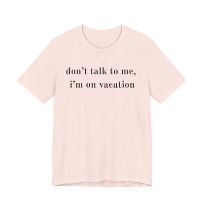 A light pink short-sleeve crewneck t-shirt with the text 'don't talk to me, i'm on vacation' printed in black serif font across the chest, displayed on a solid white background.