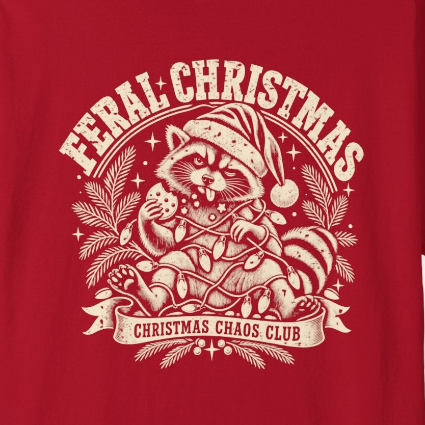 A red short-sleeve crewneck t-shirt featuring a cream-colored graphic print on the front with the text 'Feral Christmas' and an illustration of a raccoon in a Santa hat.