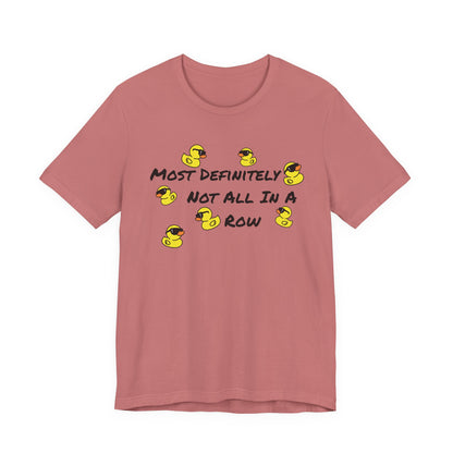 A dusty rose short-sleeve t-shirt with a graphic print on the front. The graphic features the text 'MOST DEFINITELY NOT ALL IN A ROW' in black, interspersed with small yellow rubber ducks wearing black sunglasses.