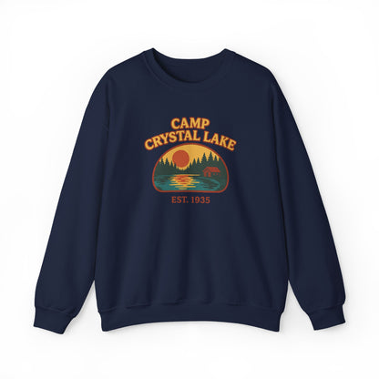 A navy blue crewneck sweatshirt featuring a retro-style 'Camp Crystal Lake' graphic on the chest with a sunset, trees, and a bear, set against a plain white background.