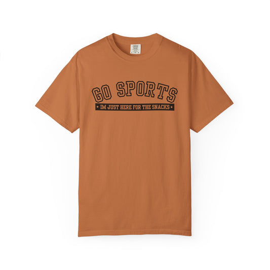 A rust-orange short-sleeve t-shirt with a black graphic on the chest that reads 'GO SPORTS' and 'I'M JUST HERE FOR THE SNACKS'.