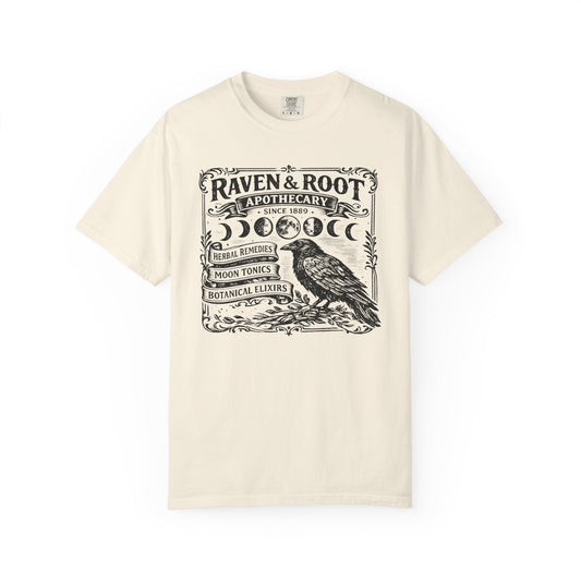 An off-white short-sleeve crewneck t-shirt featuring a black graphic print of 'Raven & Root Apothecary' with a raven illustration, moon phases, and botanical text, displayed on a clean white background.