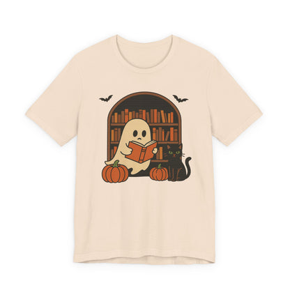 A light beige crewneck t-shirt featuring a Halloween graphic of a cute ghost reading a book in front of a bookshelf, with pumpkins, a black cat, and bats.