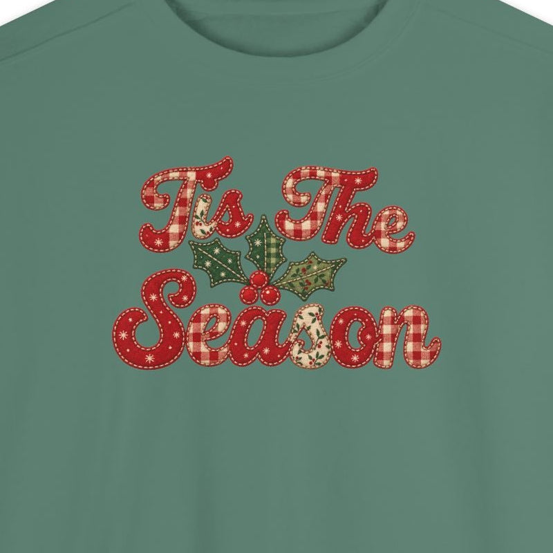 A sage green crewneck sweatshirt featuring a festive graphic on the chest that reads 'To The Season' in red and gold glittery lettering with a small green pine tree motif, displayed on a solid white background.
