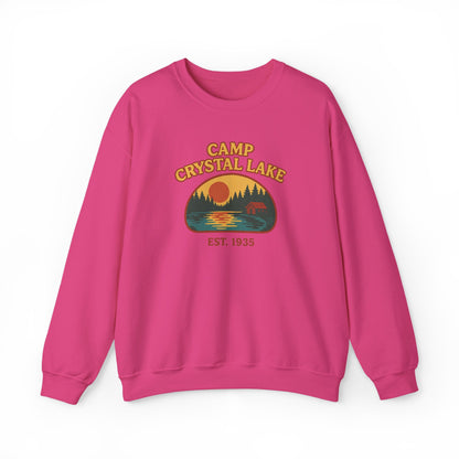 A bright pink crewneck sweatshirt featuring a retro-style 'Camp Crystal Lake' graphic on the chest with a sunset, pine trees, and a deer silhouette.