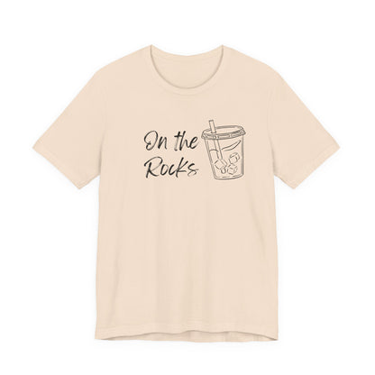 A light beige short-sleeve t-shirt with a black graphic print on the front that reads 'On the Rocks' next to a simple line drawing of a iced coffee cup, shown against a plain white background.