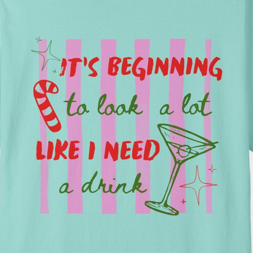 A light aqua blue short-sleeve t-shirt with a colorful graphic on the front that reads 'IT'S BEGINNING to look a lot LIKE I NEED a drink' in red and green lettering, accompanied by a martini glass and candy cane illustration, shown on a plain white background.