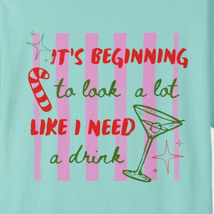 A light aqua blue short-sleeve t-shirt with a colorful graphic on the front that reads 'IT'S BEGINNING to look a lot LIKE I NEED a drink' in red and green lettering, accompanied by a martini glass and candy cane illustration, shown on a plain white background.