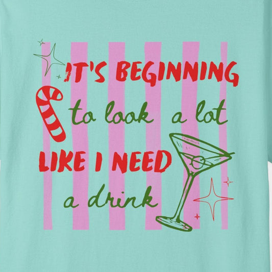 A light aqua blue short-sleeve t-shirt with a colorful graphic on the front that reads 'IT'S BEGINNING to look a lot LIKE I NEED a drink' in red and green lettering, accompanied by a martini glass and candy cane illustration, shown on a plain white background.