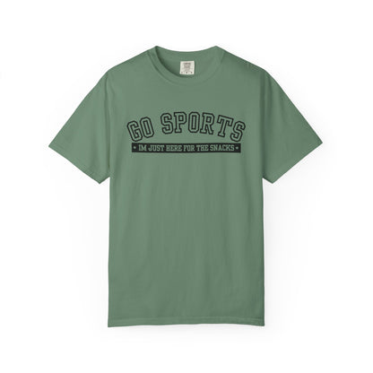 A sage green short-sleeve t-shirt with a black graphic print on the chest reading 'GO SPORTS' and 'I'M JUST HERE FOR THE SNACKS'.