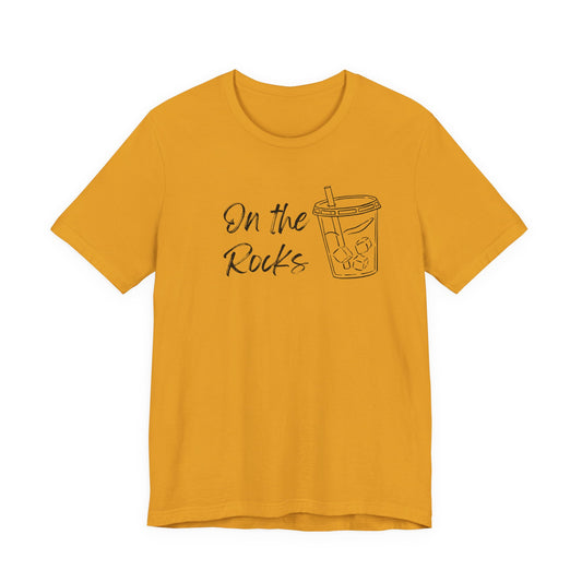 A mustard yellow short-sleeve t-shirt with a black graphic print on the front that reads 'On the Rocks' next to a simple line drawing of a bubble tea cup, shown against a plain white background.