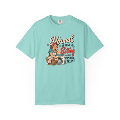A light aqua blue short-sleeve t-shirt with a vintage-style graphic on the front. The graphic features a pin-up illustration of a woman washing a shirt next to a laundry basket, with the text 'Normal is just a Setting on the Washing Machine' in stylized fonts.