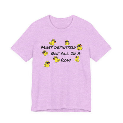 A light purple heather short-sleeve t-shirt with a graphic print on the front. The graphic features the text 'MOST DEFINITELY NOT ALL IN A ROW' in black, surrounded by small yellow rubber ducks wearing sunglasses.