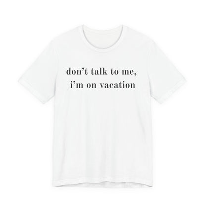A white short-sleeve crewneck t-shirt with the text 'don't talk to me, i'm on vacation' printed in black serif font across the chest, displayed on a clean white background.