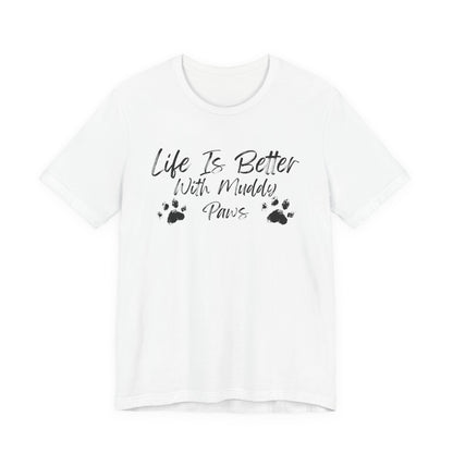 A white short-sleeve crewneck t-shirt with a black graphic print on the front that reads 'Life Is Better With Muddy Paws' alongside two small muddy paw prints, shown against a plain white background.